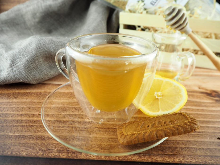 Tasse Holunderblütentee mit Zitronenscheibe und Keks auf einem Holztisch, Honiglöffel und Teepackung im Hintergrund.