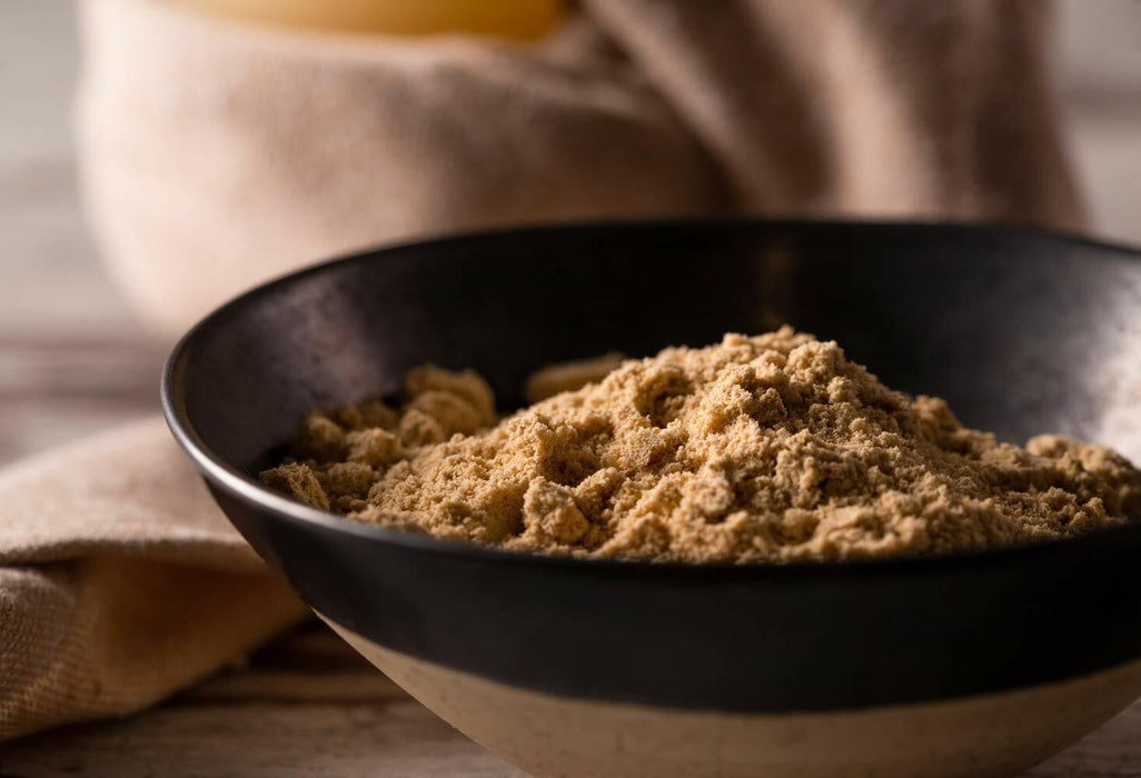 Nahaufnahme von goldenem Leinsamenpulver in einer schwarzen Schüssel, präsentiert auf einem Holztisch mit einem neutralen Stoff im Hintergrund.