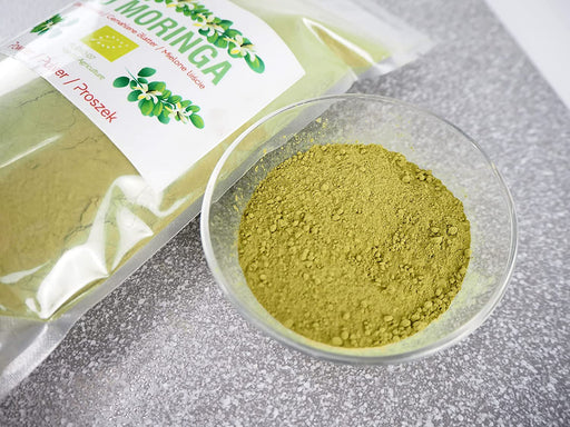 Geöffnete Packung von Bio Moringa Pulver neben einer Glasschale mit grünem Moringapulver auf einer grauen Arbeitsplatte.