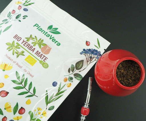 Ein rot Mate Becher gefüllt mit Yerba Mate, daneben eine Packung Bio Yerba Mate und lose Blätter auf einer dunklen Oberfläche, bereit zum Genuss.