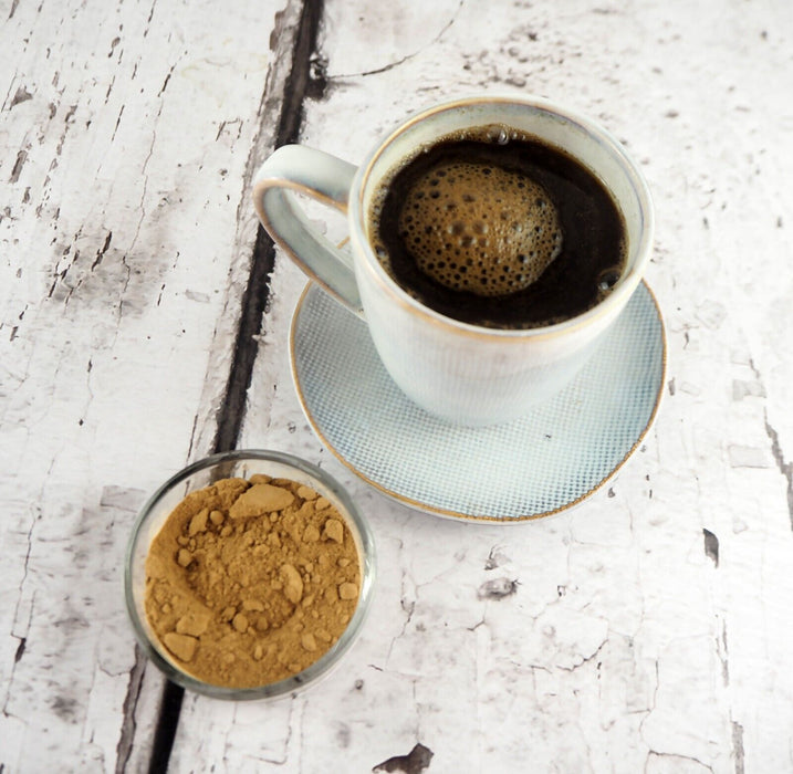 Tasse mit einem dunklen Getränk und eine Schale mit Teufelskralle-Pulver daneben, präsentiert auf einem alten weißen Holztisch.